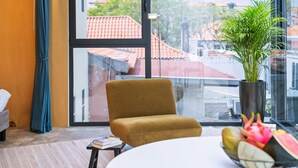 Ein Zimmer mit großem Fenster, einem Sessel, einem Tisch, einer Topfpflanze und einer Obstschale.