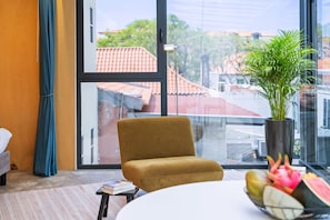 Ein Zimmer mit großem Fenster, einem Sessel, einem Tisch, einer Topfpflanze und einer Obstschale.