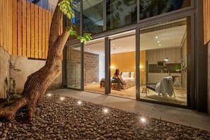 Un intérieur moderne avec une grande porte vitrée donnant sur une chambre, un coin cuisine et un petit espace extérieur agrémenté d’un arbre et de galets.