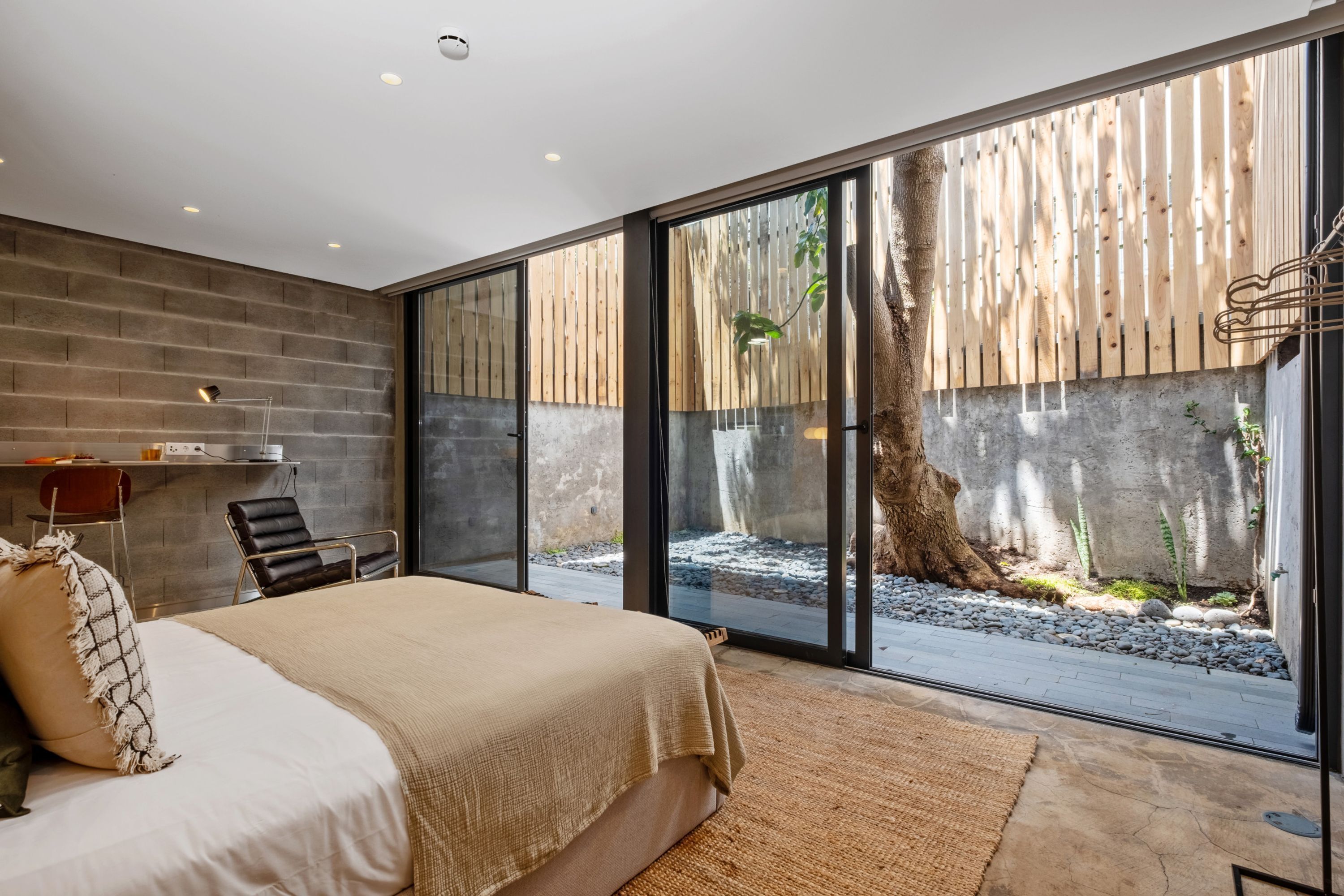 A modern bedroom with a large bed, a desk, and a view of a garden through sliding glass doors.