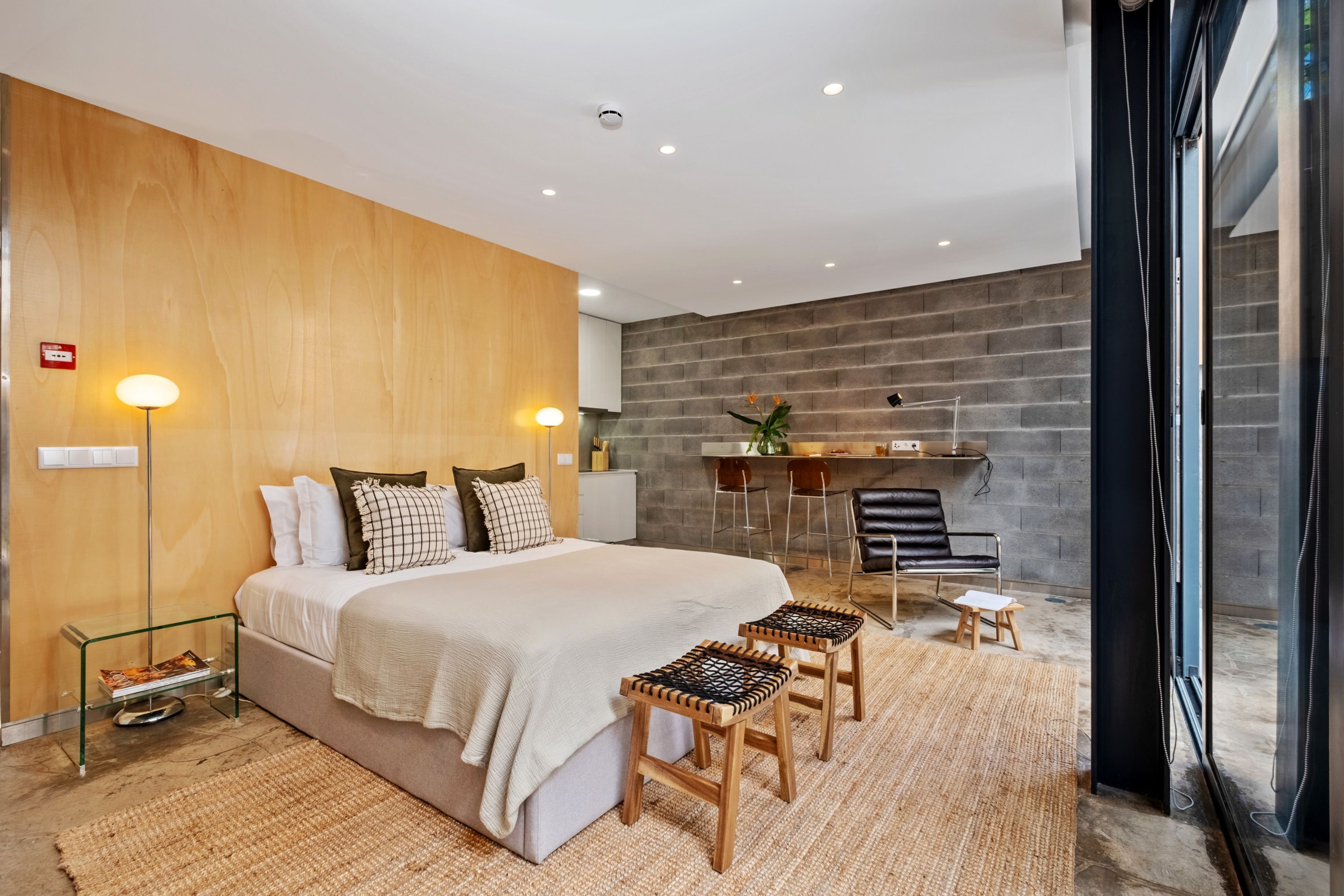 A modern bedroom with a large bed, bedside lamps, a small table, and a seating area with stools.
