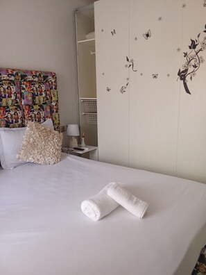 Cama bem arrumada com toalha enrolada, cabeceira colorida, espelho e parede com adesivos de flores e borboletas.