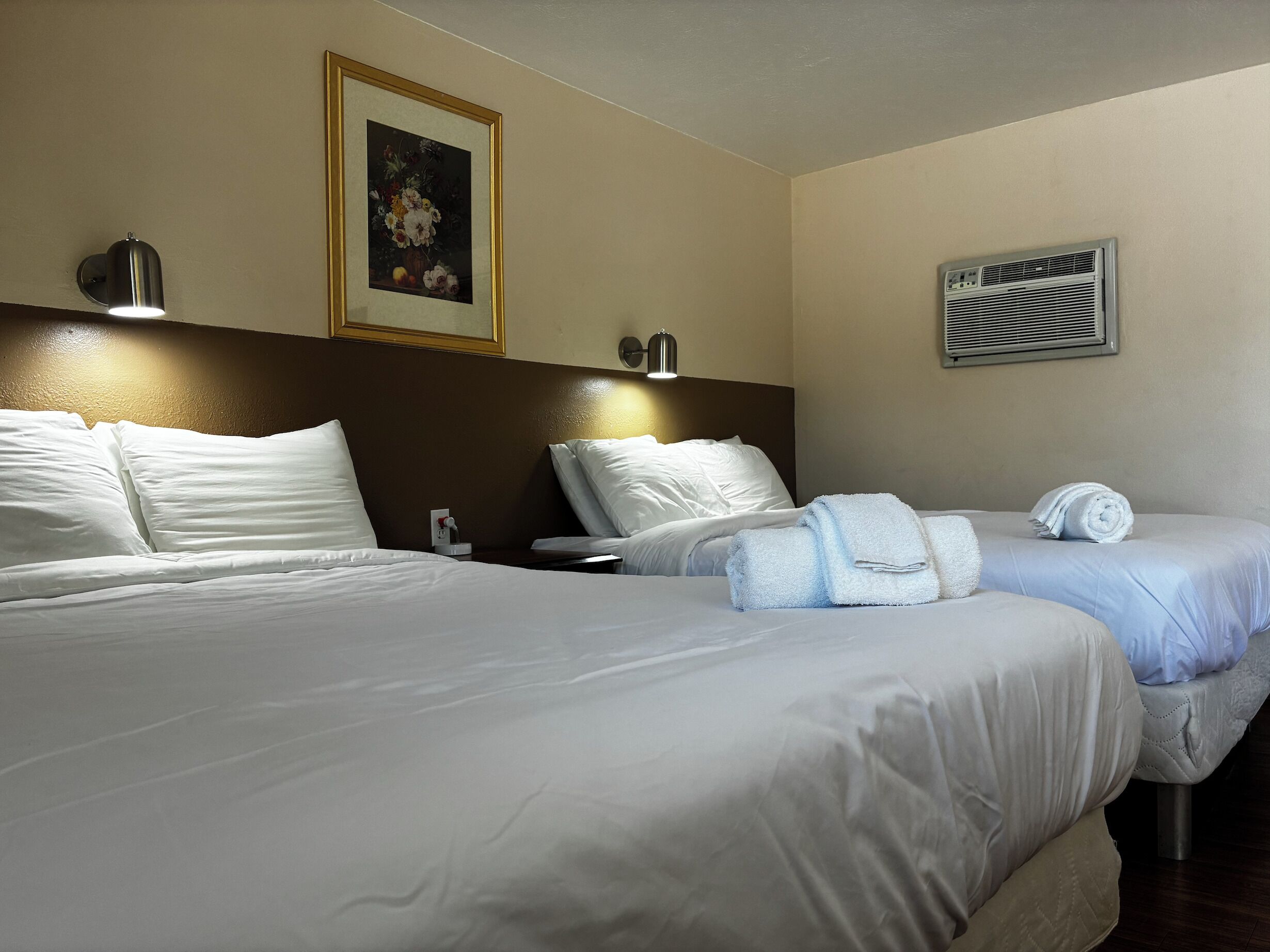 A hotel room with two beds, a framed painting, a wall-mounted air conditioner, and a bedside lamp.