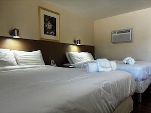 A hotel room with two beds, a framed floral painting, a wall-mounted air conditioner, and a bedside lamp.