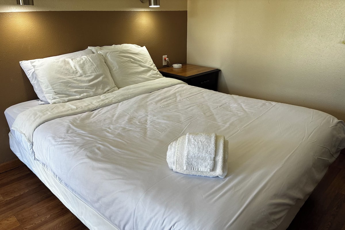 A neatly made bed with white linens, a folded white towel, and a wooden nightstand with a lamp.