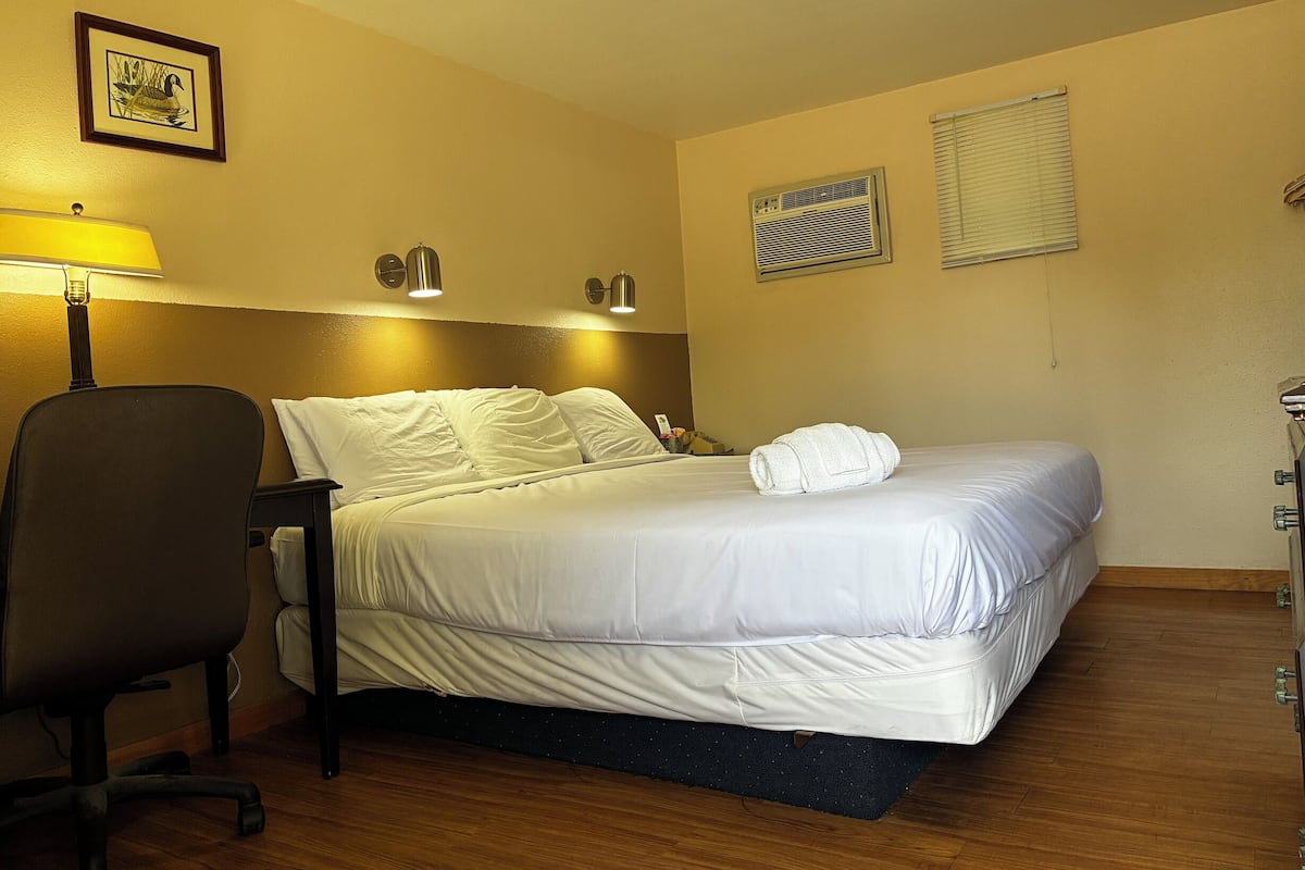 A hotel room with a bed, a desk, a chair, a lamp, two wall-mounted lights, a framed picture, an air conditioner, and a window with blinds.