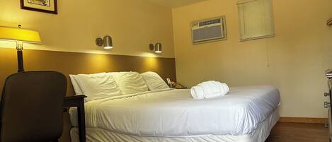 A hotel room with a bed, a desk, a chair, a lamp, two wall-mounted lights, a framed picture, an air conditioner, and a window with blinds.