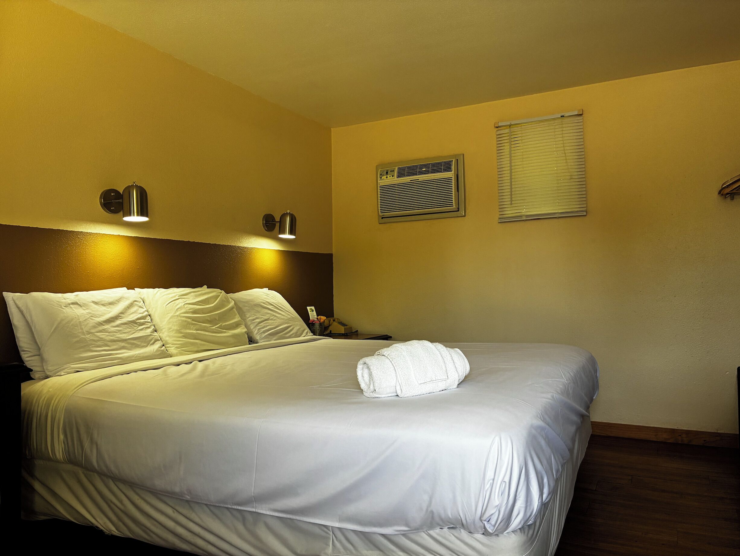 Une chambre d’hôtel comprenant un lit, deux lampes fixées au mur, une fenêtre avec des stores et un climatiseur fixé au mur.
