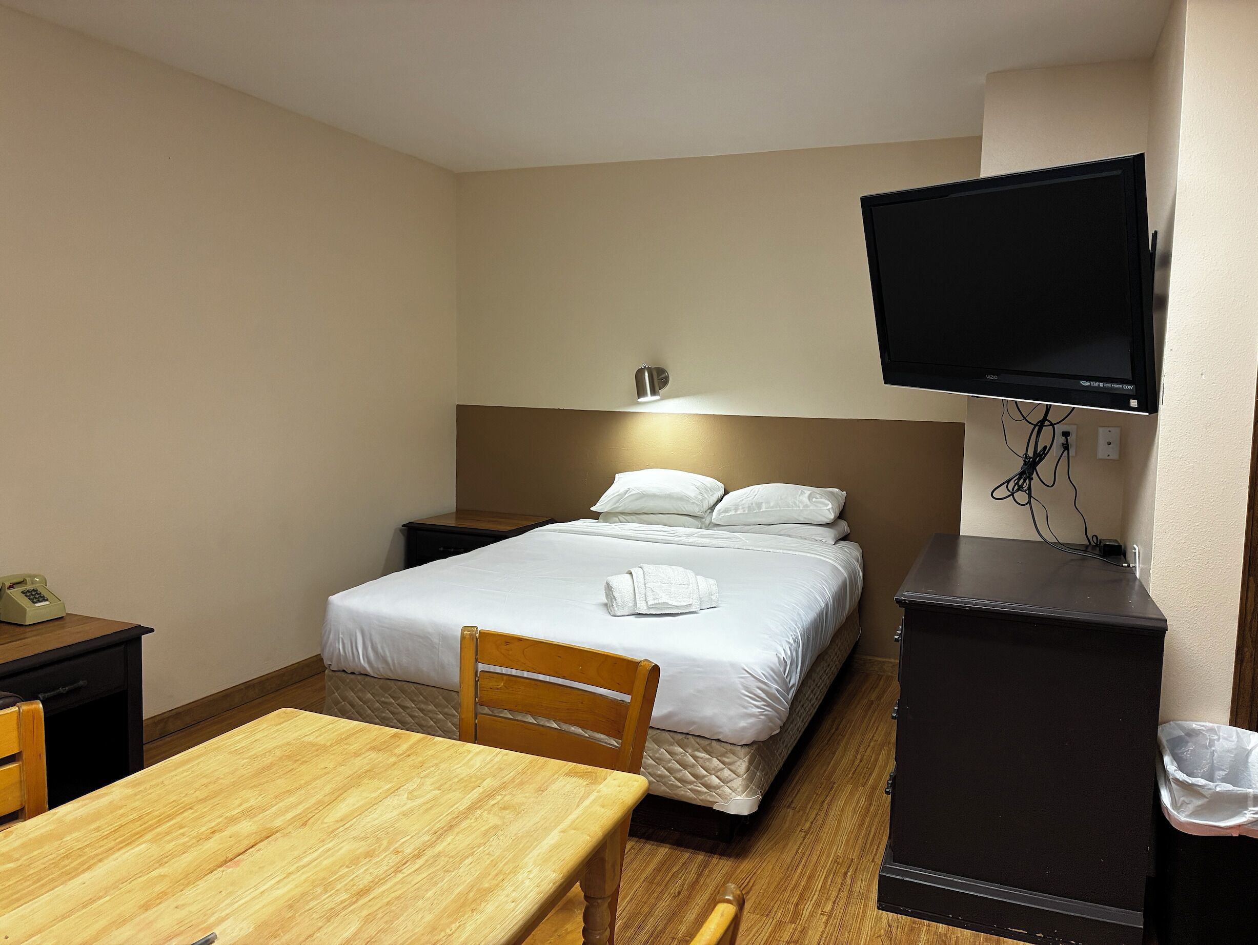 A hotel room with a bed, bedside table, chair, desk, TV, and a trash bin.