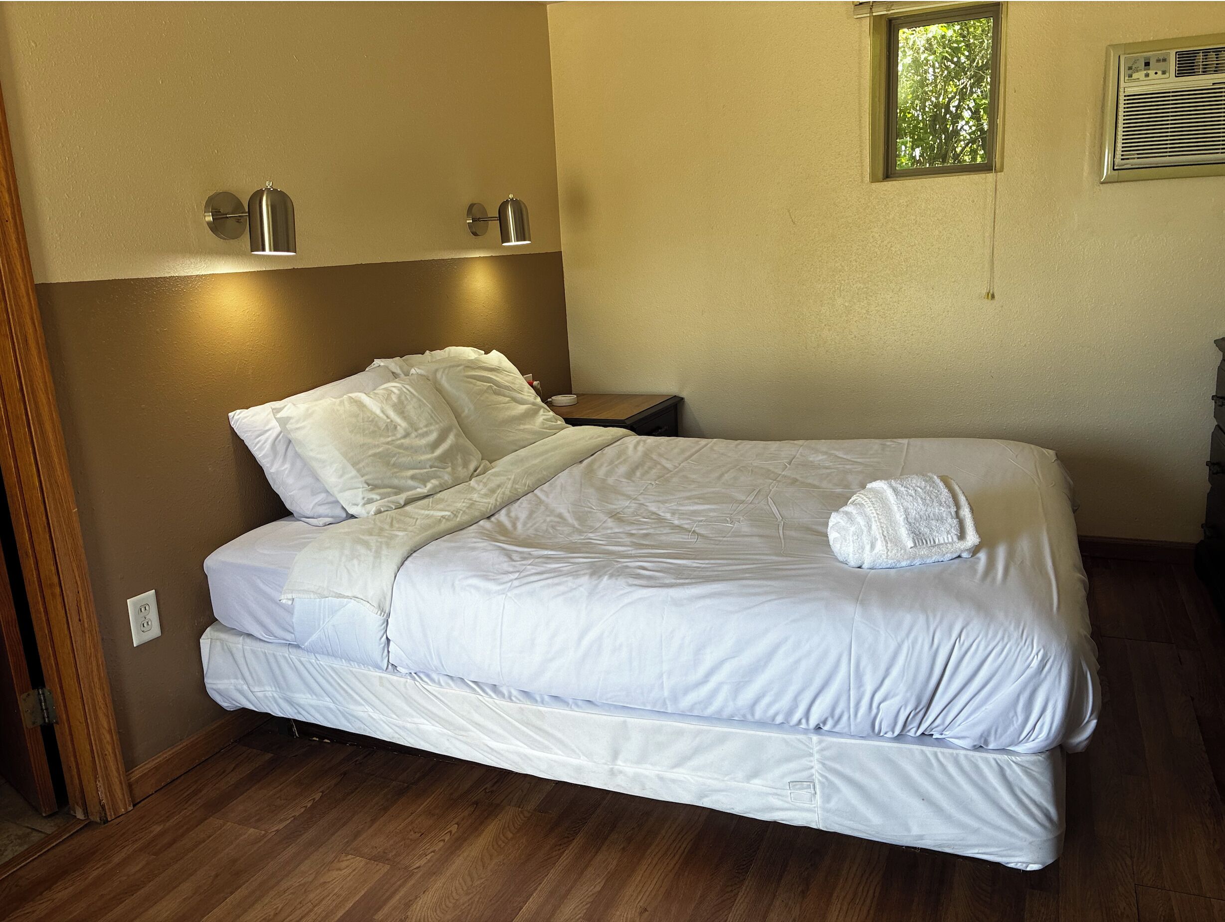 A neatly made bed with white linens, a wooden floor, and two wall-mounted lamps.