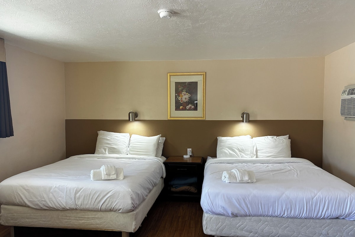 A hotel room with two beds, a painting on the wall, and a ceiling-mounted air conditioning unit.