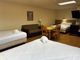 Habitación de hotel con dos camas individuales, un sofá, una mesa de comedor con sillas, un televisor y una lámpara.