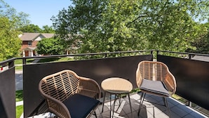 Een balkon met rieten stoelen en een tafeltje, met uitzicht op een woonwijk met bomen en huizen.