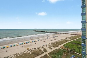 A beach with a pier extending into the water, a sandy beach area with umbrellas, and a building with a view of the beach. - Oceanfront King Room + Lazy River & Hot Tub (Myrtle Beach)