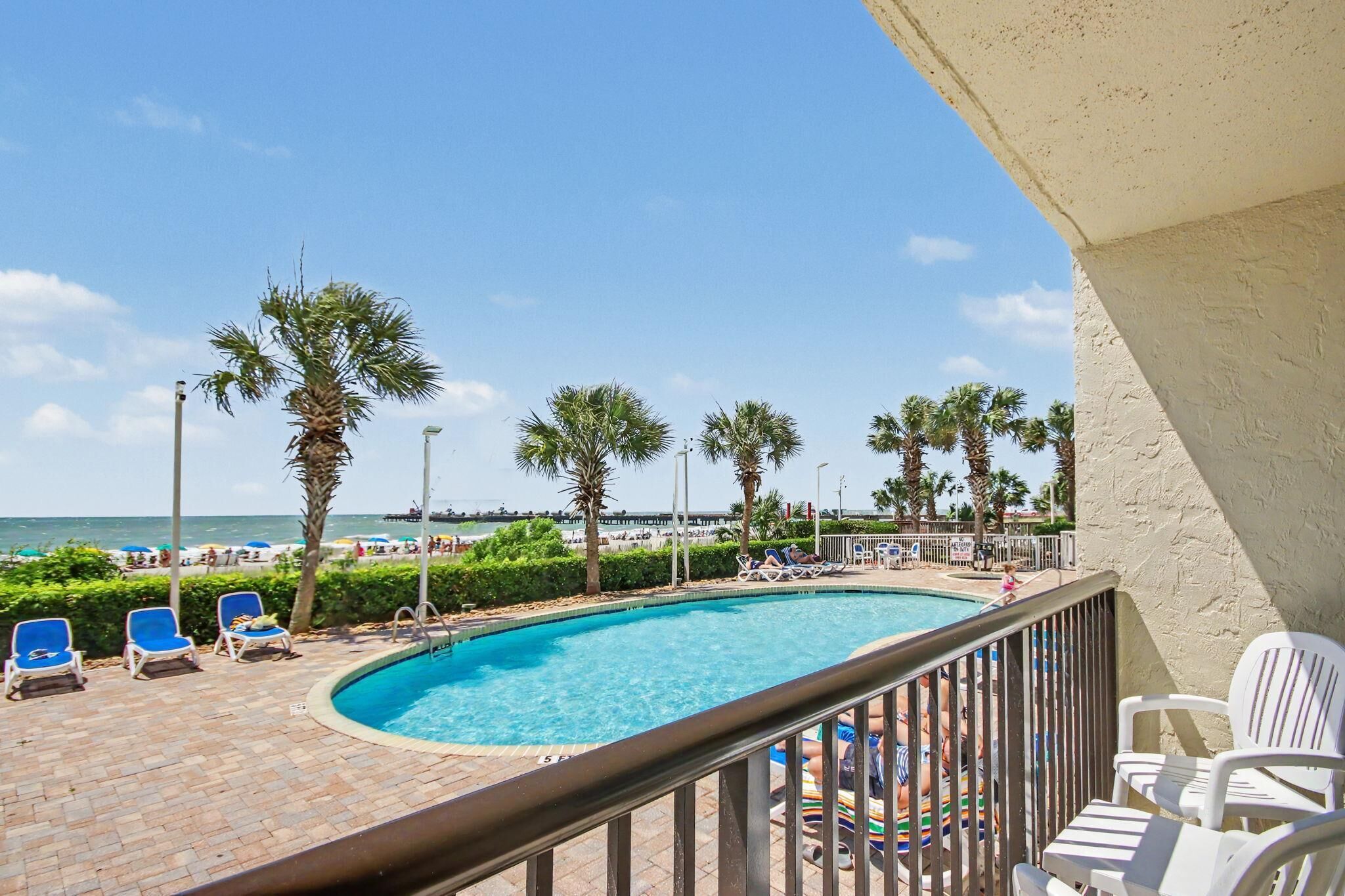 A pool area with lounge chairs, a clear view of the beach, and a distant pier.
