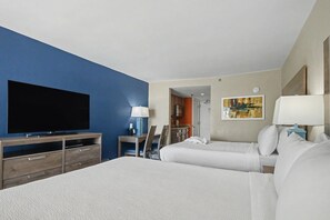 A hotel room with a large flat-screen TV, a bed with white linens, a desk with a lamp, and a painting on the wall. - Oceanfront Two Queen Room + Lazy River & Hot Tub (Myrtle Beach)