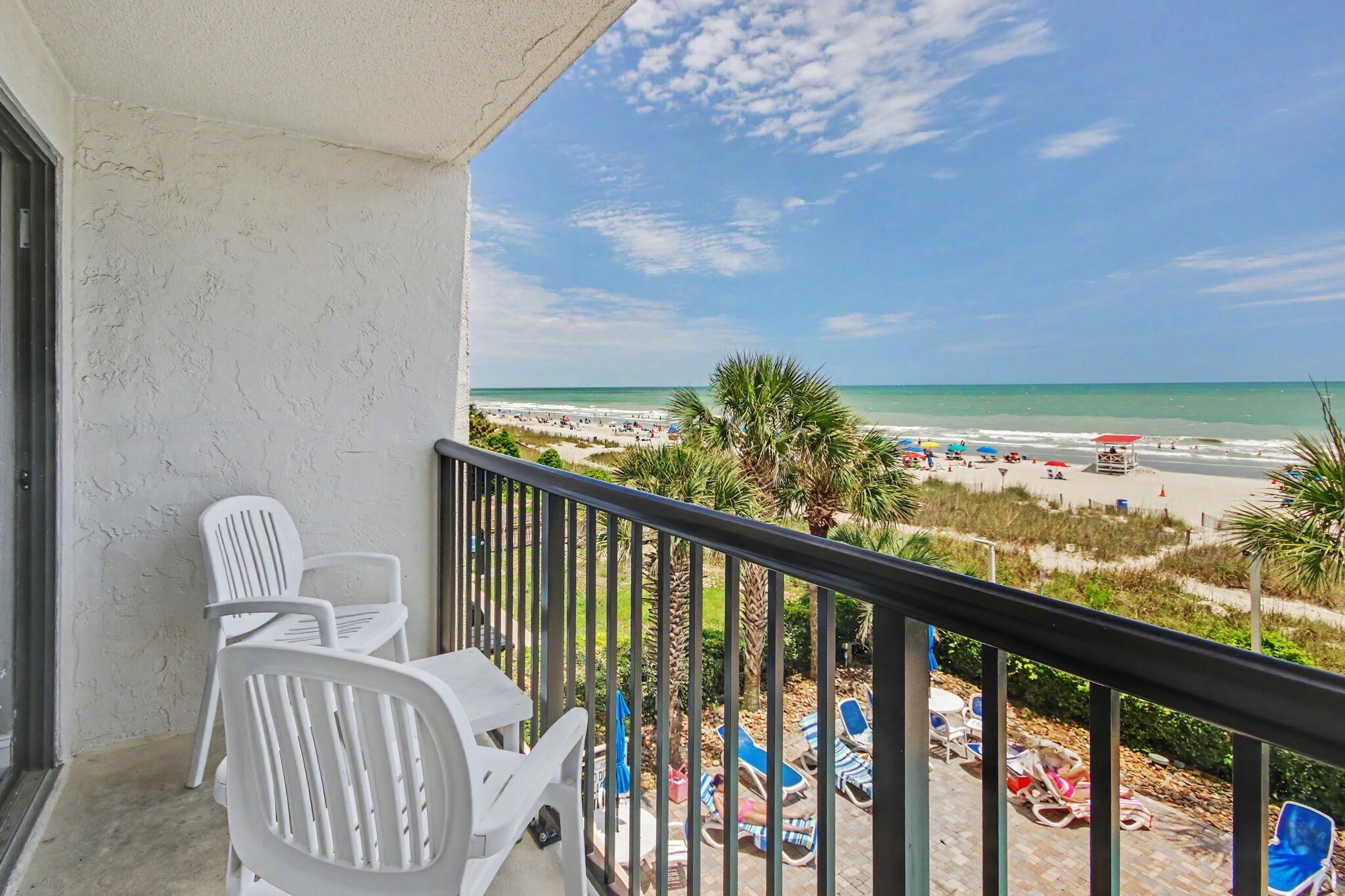 A balcony with white plastic chairs, a view of a beach with umbrellas and people, and a clear sky.
