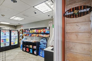 A snack bar with shelves of snacks, a freezer, and a counter with a sign reading 'The Snack Shack'. - Oceanfront Two Queen Room + Lazy River & Hot Tub (Myrtle Beach)