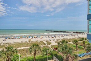 A beach with a pier extending into the water, palm trees, and a building with a balcony overlooking the beach. - Oceanfront Two Queen Room + Lazy River & Hot Tub (Myrtle Beach)