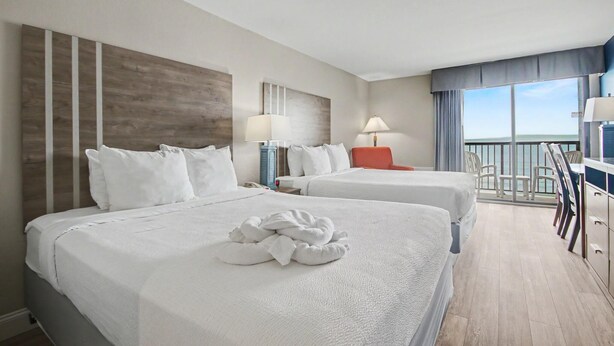A hotel room with two beds, a balcony with chairs, and a view of the sea. - Oceanfront Two Queen Room + Lazy River & Hot Tub (Myrtle Beach)