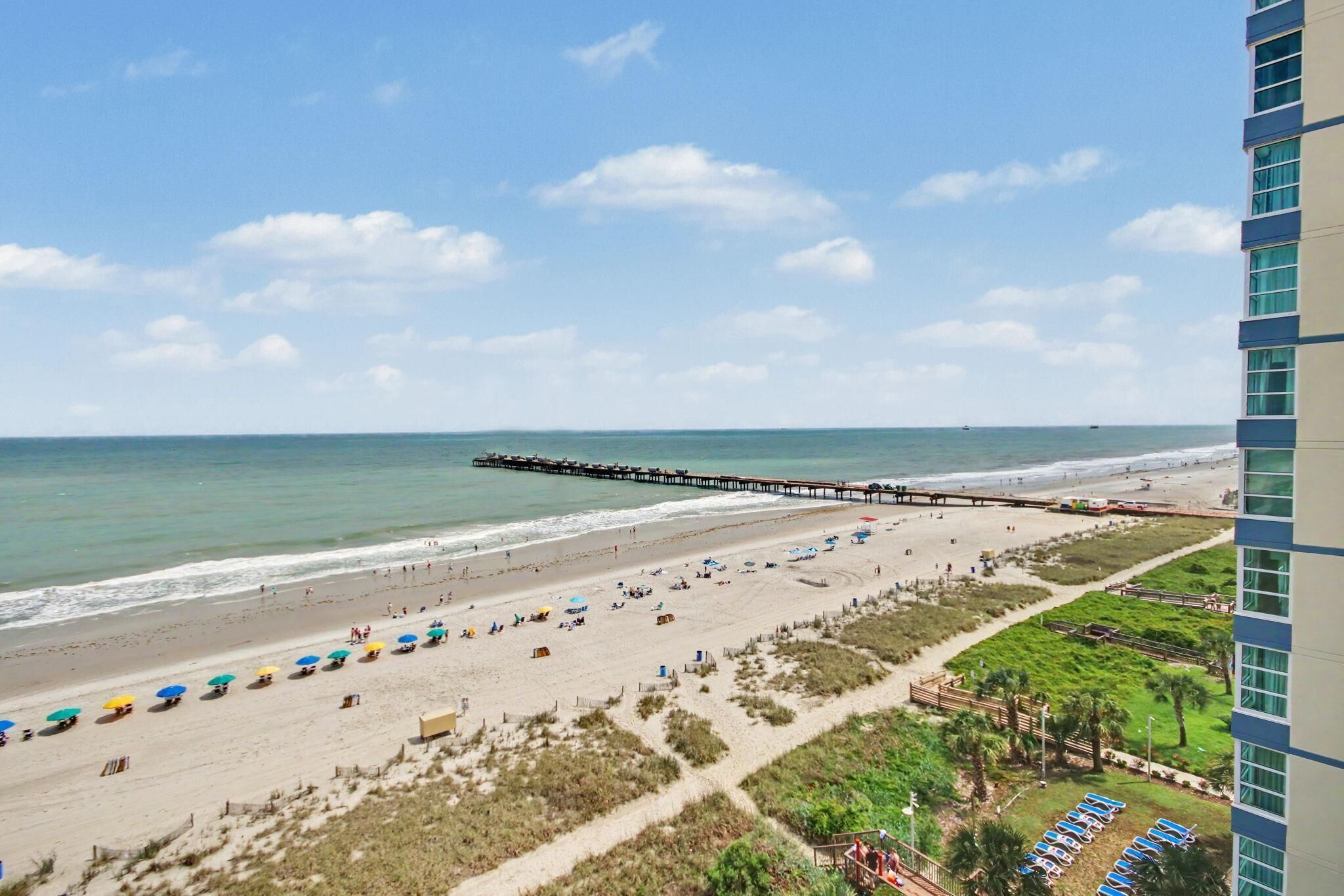 Ein Strand mit einem Steg, der sich ins Meer erstreckt, Sand und Sonnenschirme sowie ein Gebäude mit großen Fenstern.