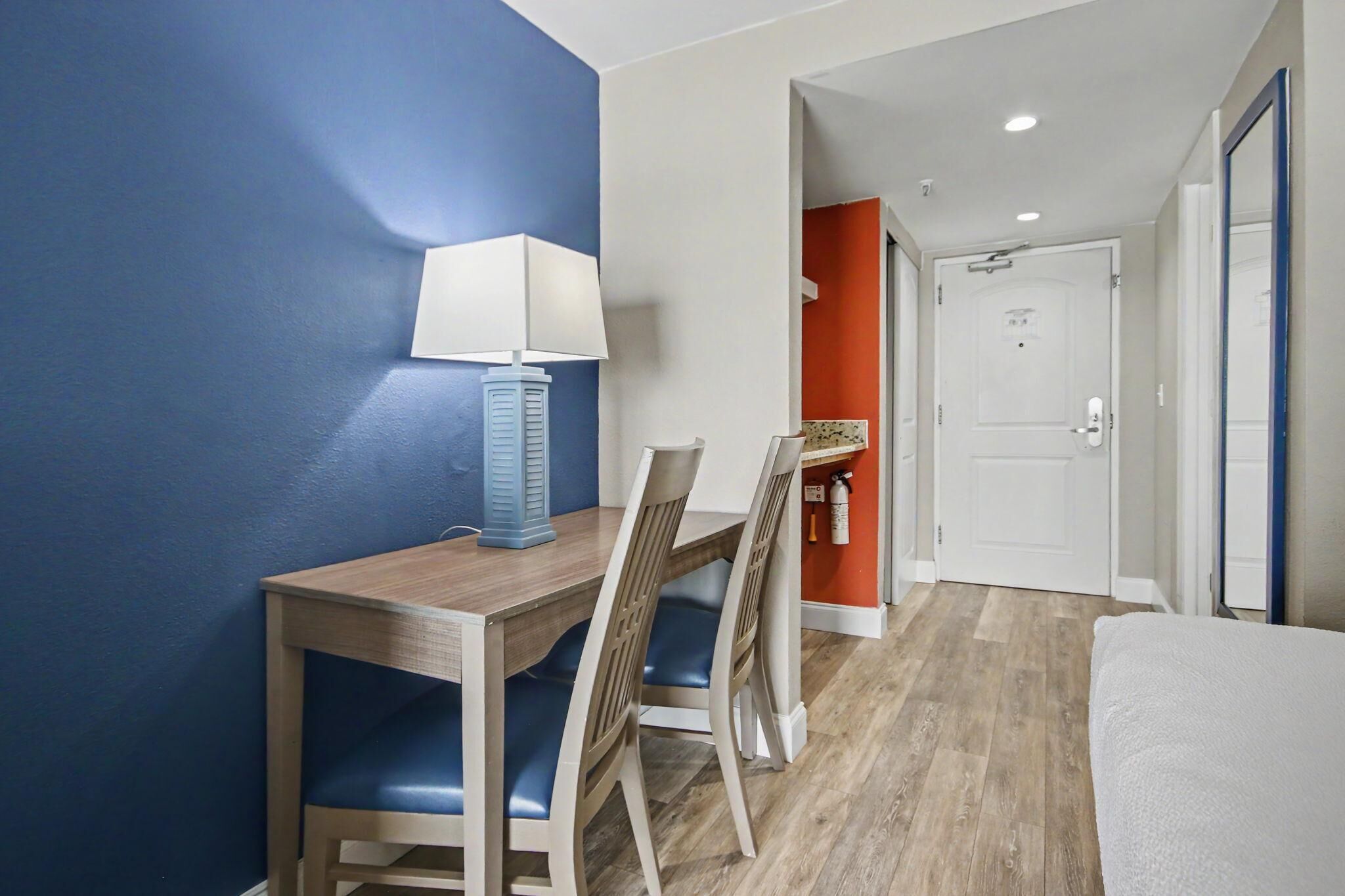 Une chambre d’hôtel avec un bureau en bois, des chaises bleues, un lit blanc et une porte donnant sur une autre pièce.