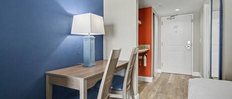 Une chambre d’hôtel avec un bureau en bois, des chaises bleues, un lit blanc et une porte donnant sur une autre pièce.