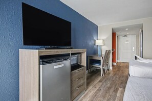 A compact hotel room with a flat-screen TV mounted above a built-in dishwasher and oven, a small desk with a chair, a bed, and a door leading to another room. - Oceanfront Two Queen Standard Room + Lazy River & Hot Tub (Myrtle Beach)