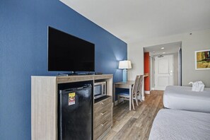 A hotel room with a flat-screen TV, a mini-fridge, a desk with a chair, a bed, and a door leading to another room. - Oceanfront Two Queen Standard Room + Lazy River & Hot Tub (Myrtle Beach)