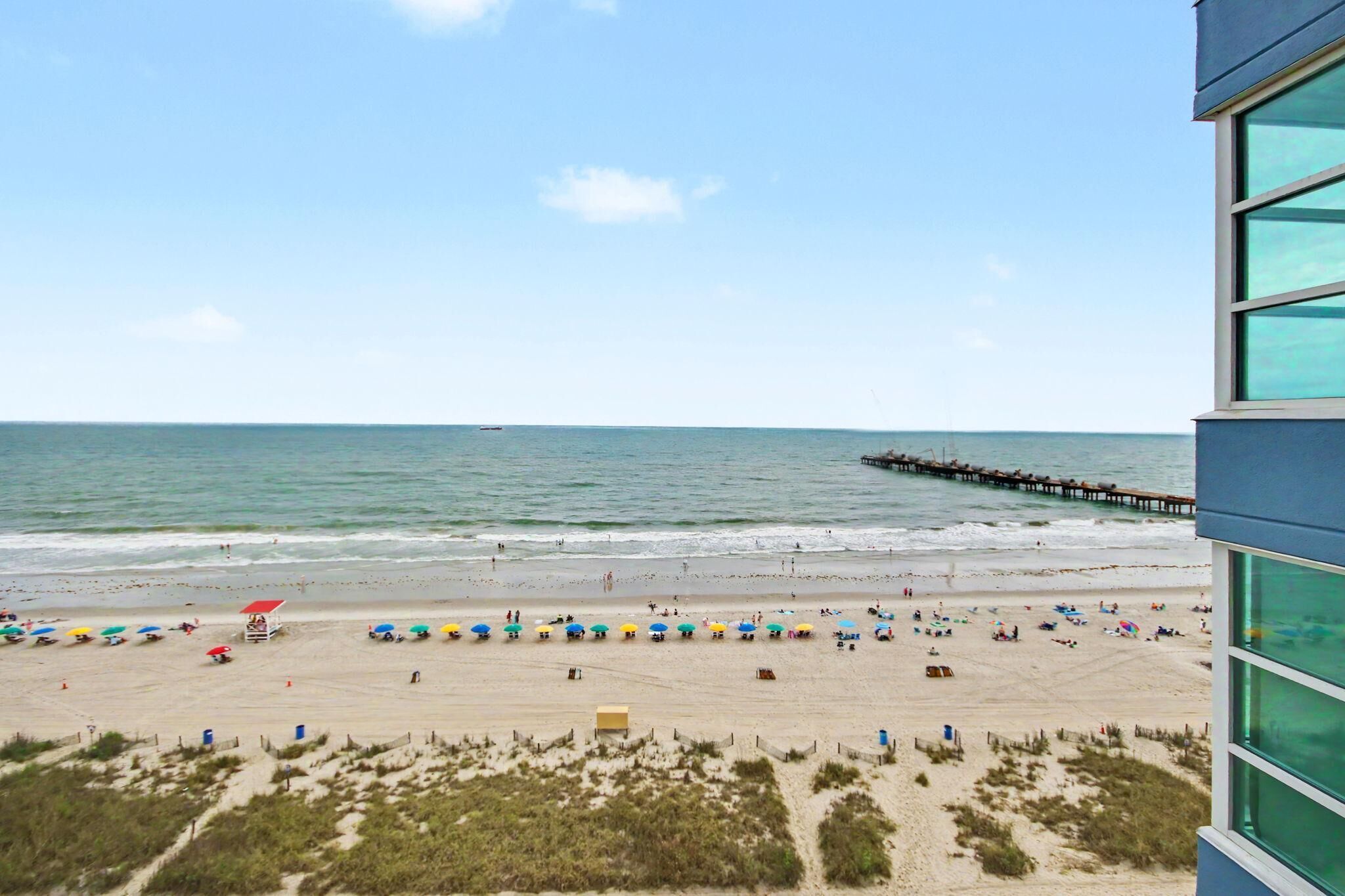 A beach with umbrellas and people, a pier extending into the water, and a building with large windows.