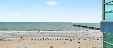 A beach with umbrellas and people, a pier extending into the water, and a building with large windows.