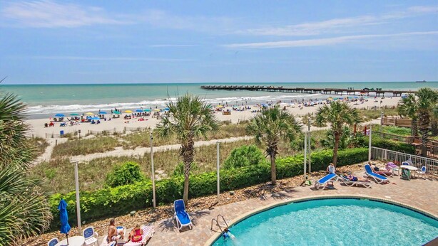 A beach resort with a swimming pool, beach umbrellas, lounge chairs, and a pier extending into the ocean. - Oceanfront Two Queen Room + Lazy River & Hot Tub (Myrtle Beach)