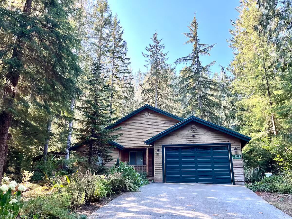 Une maison en bois d’un seul étage, avec une porte de garage sombre, entourée d’une forêt dense.
