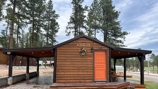 A wooden cabin with a sign reading 'Bear's Den,' a covered porch, and a small outdoor seating area. - Bears Den, studio cabin, full kitchen, cover carport, Restmore Inn, Hill City (Hill City)