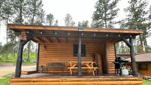 A wooden cabin with a covered porch, a bench, a table, and a grill. - Bears Den, studio cabin, full kitchen, cover carport, Restmore Inn, Hill City (Hill City)