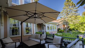 A deck with outdoor furniture, a large umbrella, and a view of a garden with flowers and a tree. - The Villa Callo Inn (West Harwich)