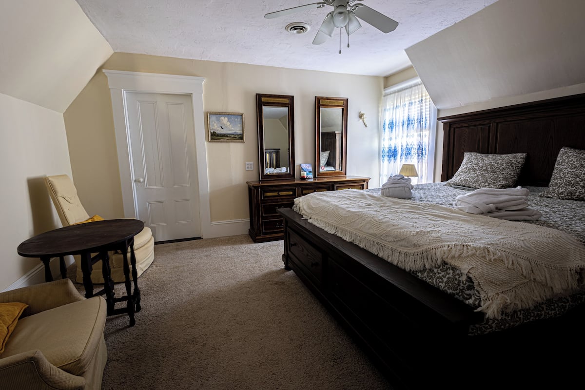 A bedroom with a bed, a chair, a table, a dresser, and a ceiling fan.