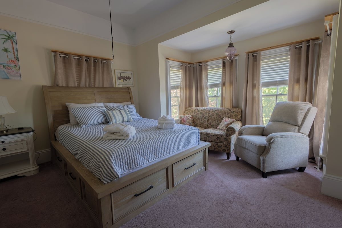 A bedroom with a bed, two chairs, a nightstand, a lamp, and a window with curtains.