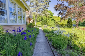 A pathway flanked by vibrant flowers and greenery, leading to a house with large windows.