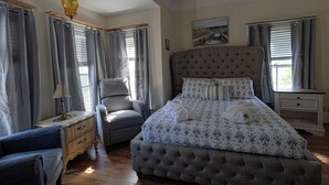 A bedroom with a large bed, a chair, a nightstand, and a window with curtains. - The Villa Callo Inn (West Harwich)
