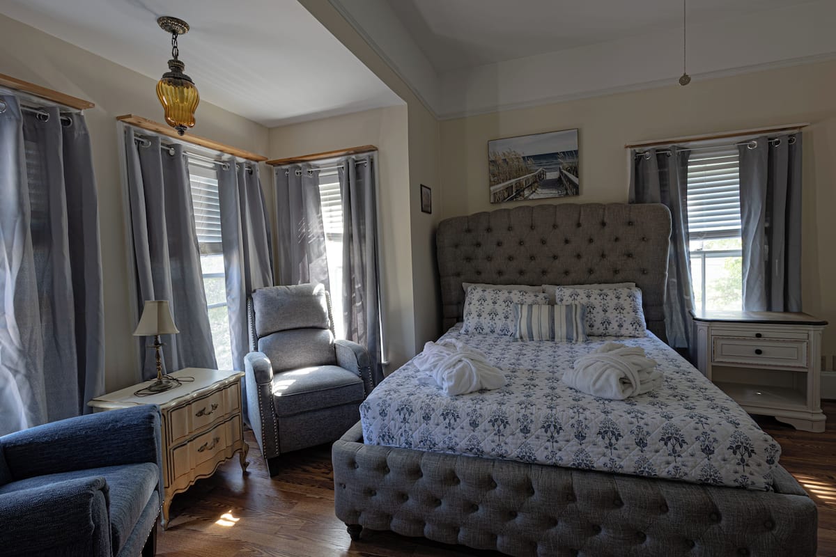A bedroom with a large bed, a chair, a nightstand, and a window with curtains.