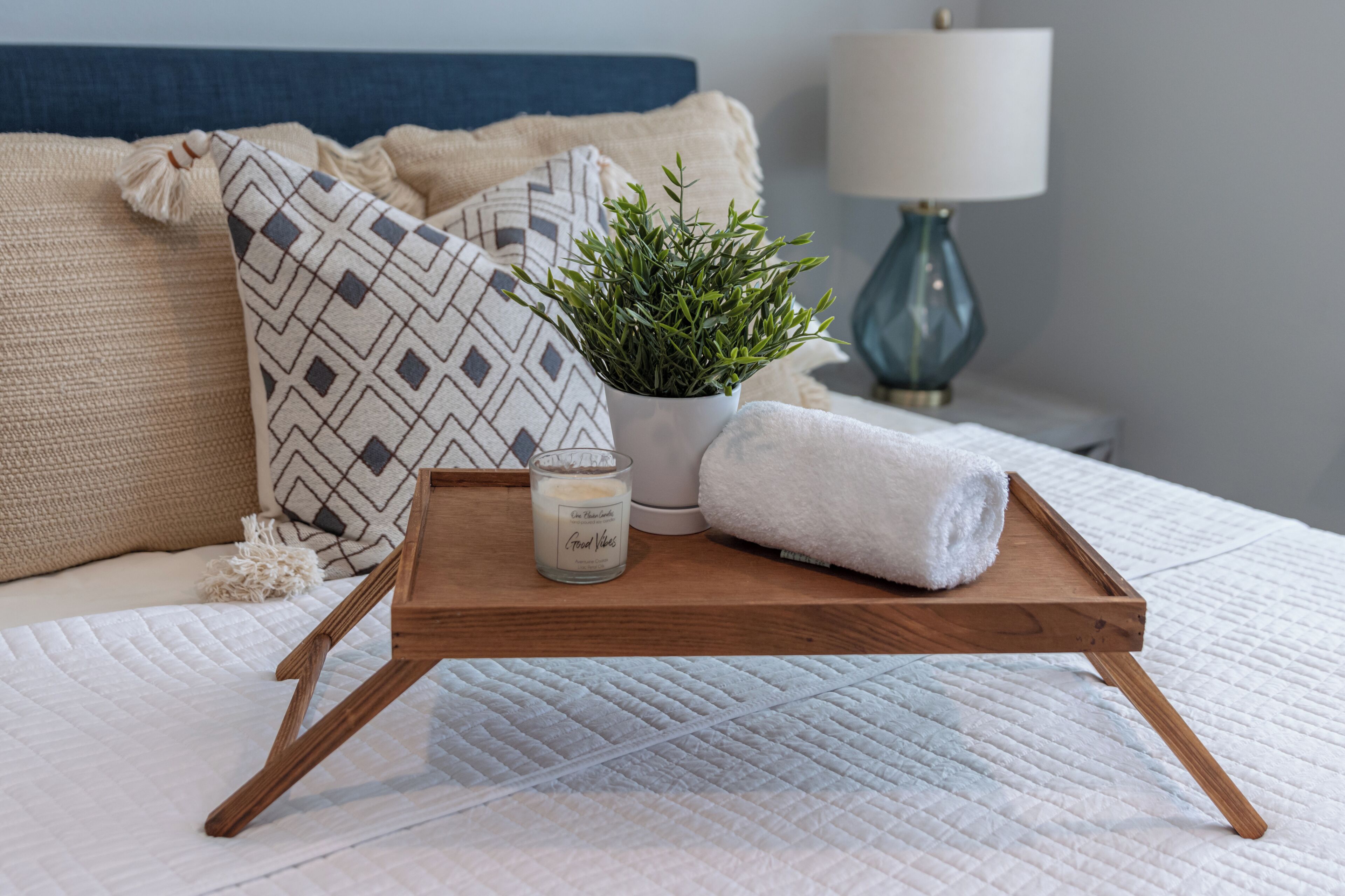 A neatly arranged bedroom with a bed, a bedside table with a lamp, a potted plant, a candle, and a tray with a towel.