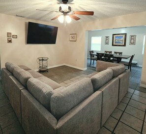 A living room with a sectional sofa, a ceiling fan, a flat-screen TV, a dining area with a table and chairs, and a tiled floor.