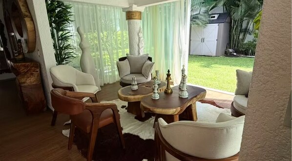 Ein Wohnzimmer mit einer Couch, Sesseln, einem Couchtisch und Blick auf einen Garten mit Rasen und Palmen.