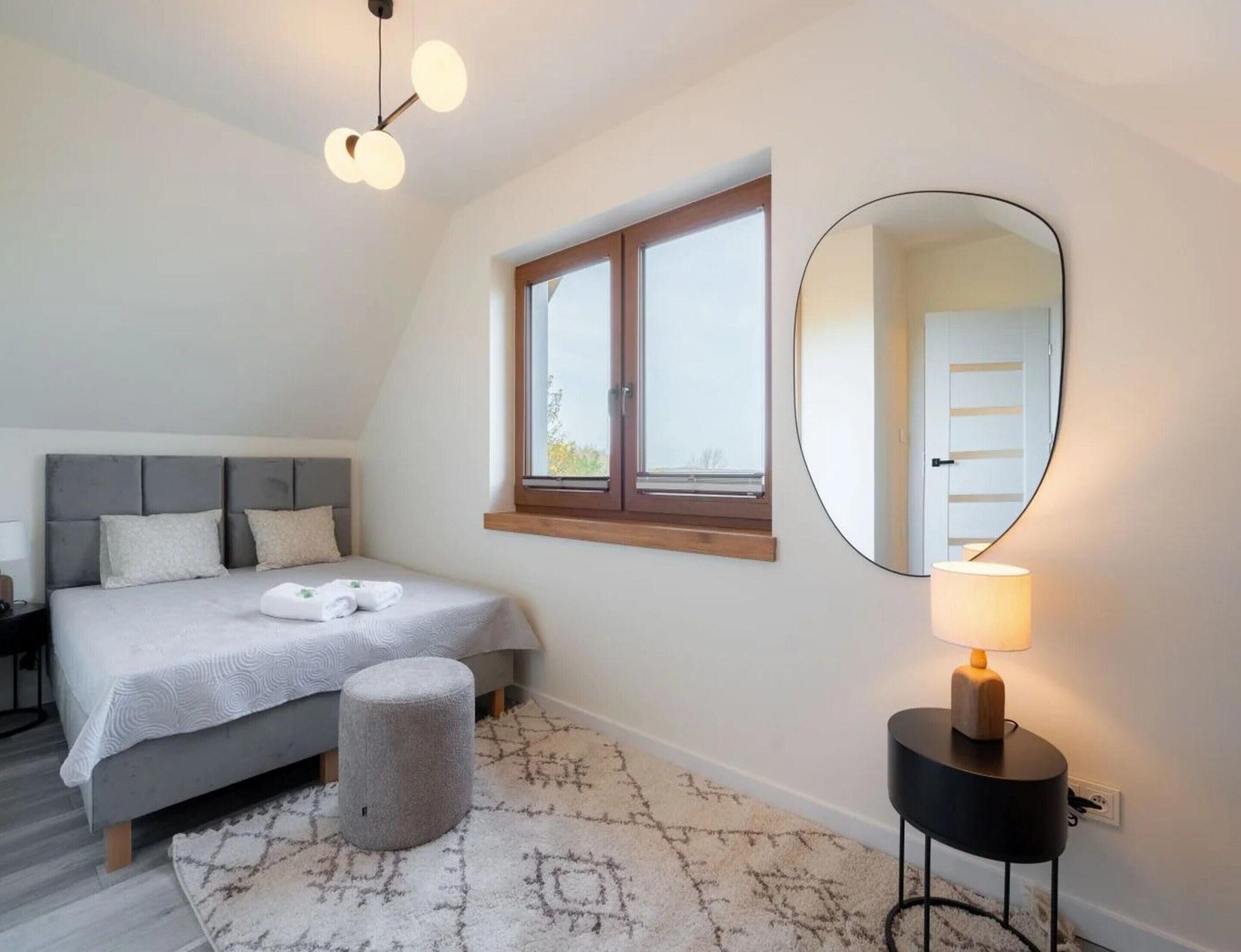 Un dormitorio con cama, espejo redondo, mesita de noche, lámpara y ventana.