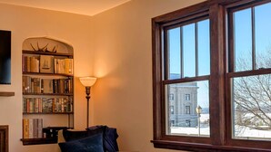 Une pièce avec une bibliothèque, une lampe, une chaise et une fenêtre donnant sur un bâtiment à l’extérieur.