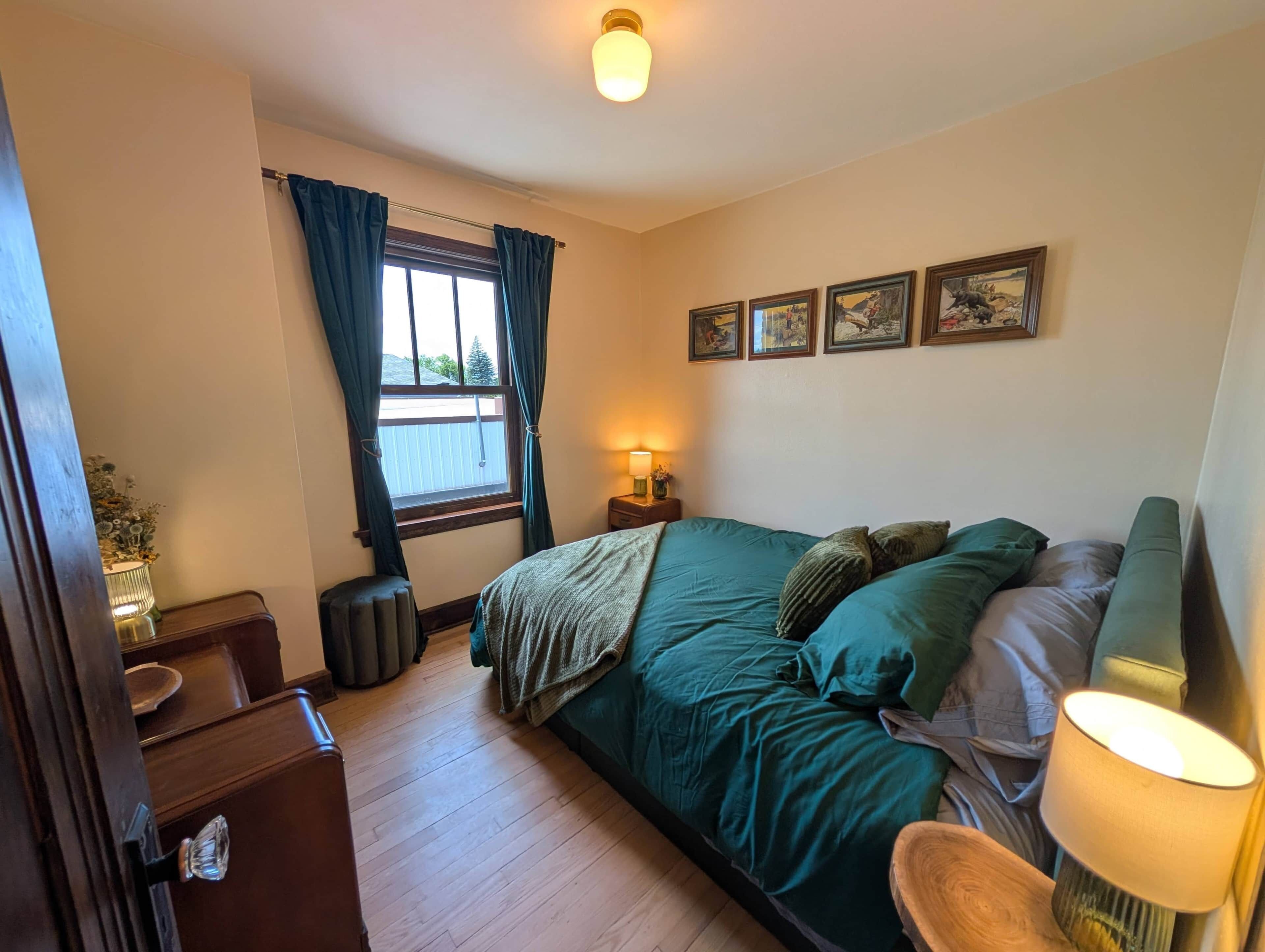 Een slaapkamer met een bed, een raam met gordijnen, een bedlamp, een klein tafeltje en een aan de muur hangend schilderij.