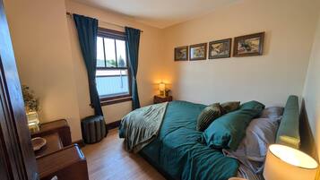 Een slaapkamer met een bed, een raam met gordijnen, een bedlamp, een klein tafeltje en een aan de muur hangend schilderij.