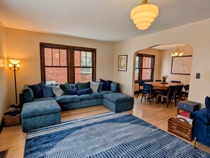 A living room with a sectional sofa, a dining table with chairs, and a large window with a view of a brick building. - Historic Main St w/ Lake View | The Flatwater Loft (Ashland)