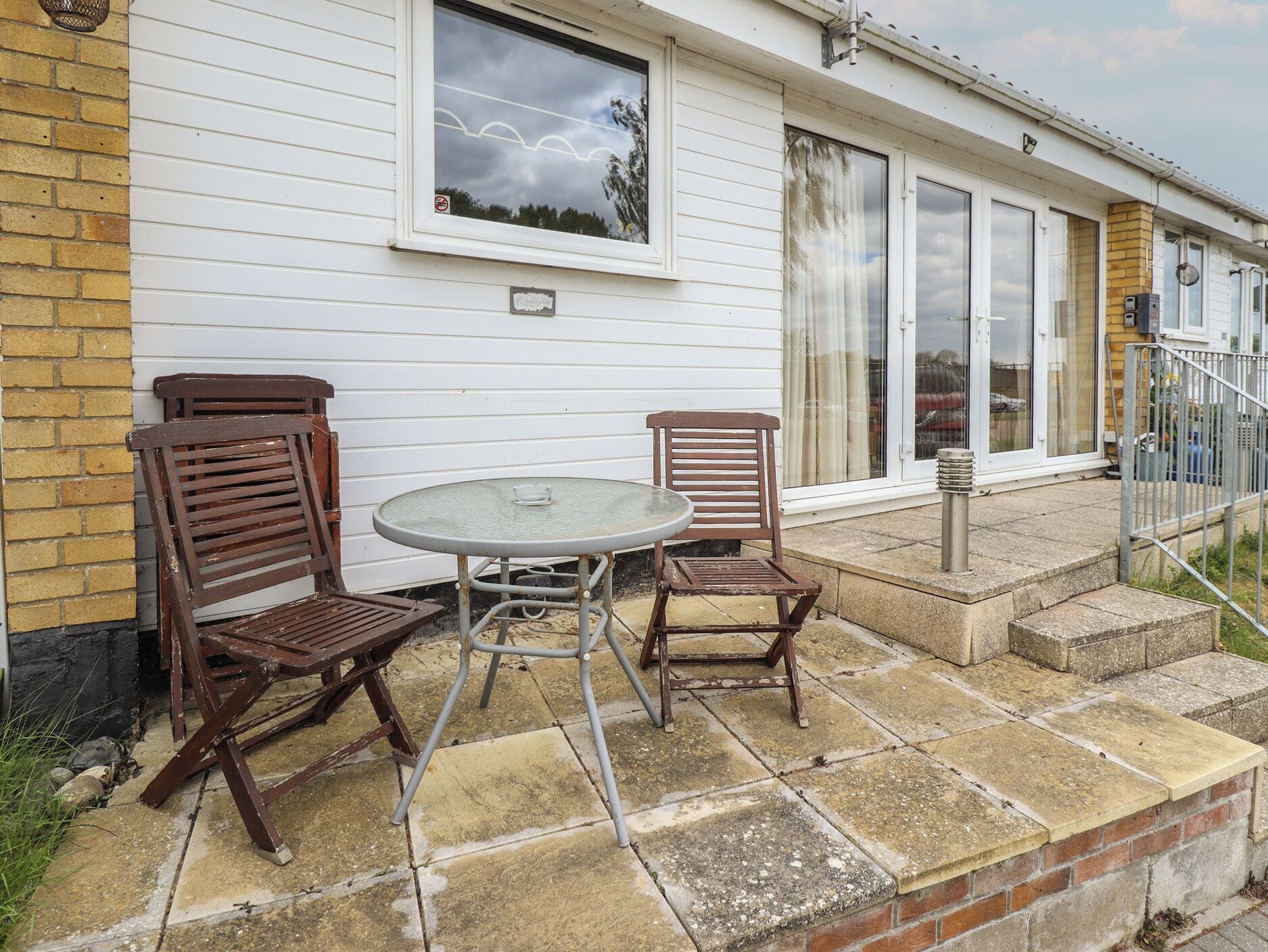 A patio with two wooden chairs and a glass table, a brick wall, and a white building with large windows and a door.